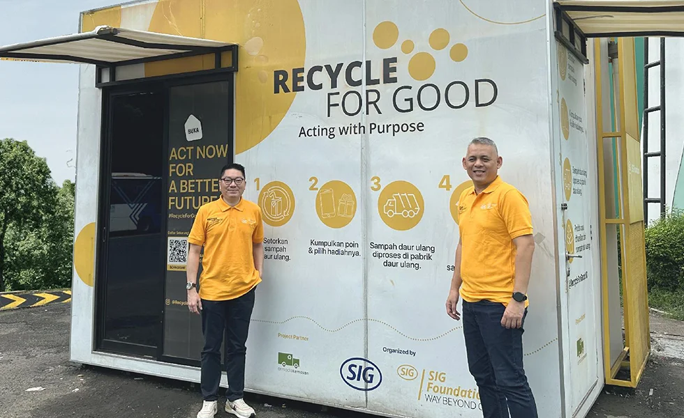 The image features two men standing in front of a recycling center or a small yellow building, which has 